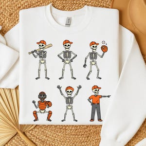 May include: White sweatshirt featuring six cartoon skeletons in baseball poses. Each skeleton wears an orange baseball cap and is engaged in different baseball activities, such as batting, pitching, and catching.