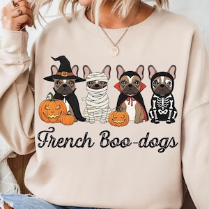 May include: Beige sweatshirt featuring four French Bulldogs in Halloween costumes: a witch, a mummy, a vampire, and a skeleton. Each dog is next to a carved pumpkin. The text "French Boo-dogs" is printed below the dogs.