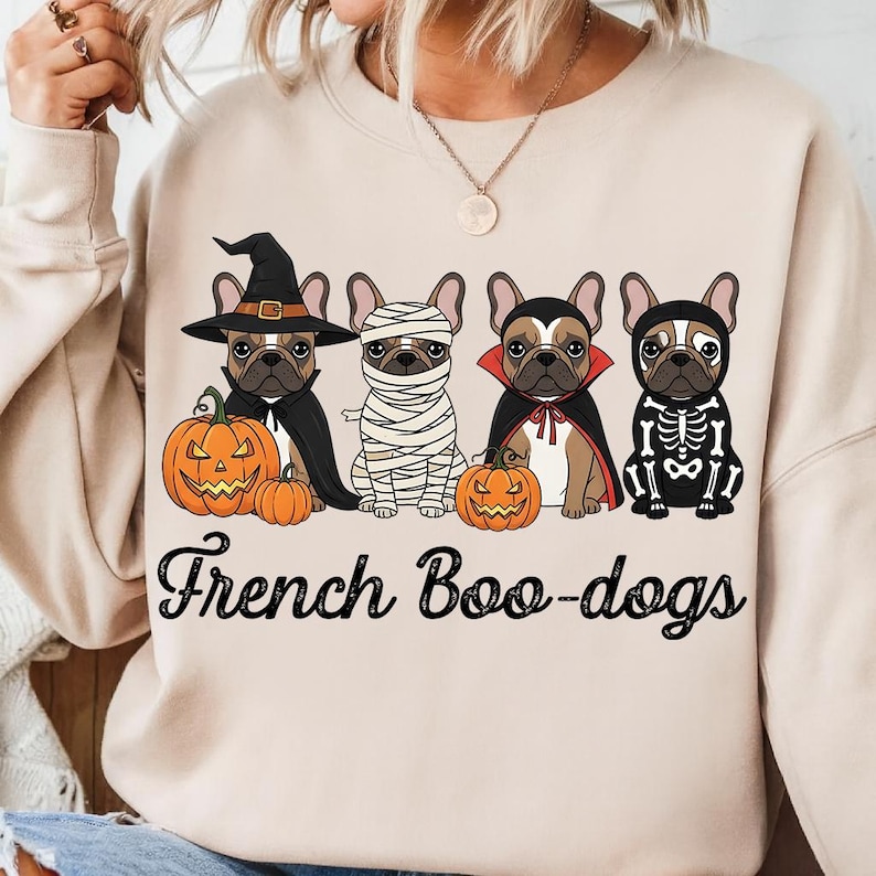 May include: Beige sweatshirt featuring four French Bulldogs in Halloween costumes: a witch, a mummy, a vampire, and a skeleton. Each dog is next to a carved pumpkin. The text "French Boo-dogs" is printed below the dogs.
