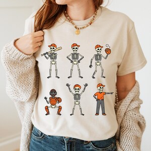 May include: Beige t-shirt featuring a baseball-themed skeleton design. The skeletons are depicted in various baseball positions, wearing orange caps and holding baseball bats, gloves, and balls. The design is a fun and unique take on the sport.