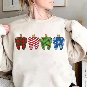 Puede incluir: Una sudadera color crema con cuatro adornos navide&ntilde;os en forma de diente. Los adornos est&aacute;n decorados con un patr&oacute;n de cuadros rojos, rayas rojas y blancas, acebo verde y copos de nieve azules. Cada adorno tiene una tapa dorada.