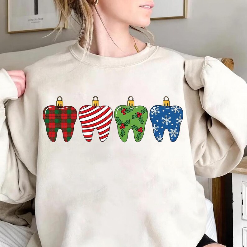 Puede incluir: Una sudadera color crema con cuatro adornos navide&ntilde;os en forma de diente. Los adornos est&aacute;n decorados con un patr&oacute;n de cuadros rojos, rayas rojas y blancas, acebo verde y copos de nieve azules. Cada adorno tiene una tapa dorada.