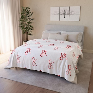 May include: White throw blanket with a red bow pattern. The blanket is on a bed with white pillows.