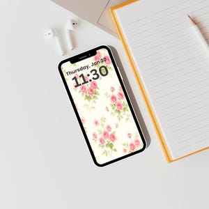 May include: A white smartphone displays a floral wallpaper of pink roses and green leaves against a cream background. The screen shows the date and time: Thursday, Jan 30, 11:30. White earbuds and a yellow notebook with a pencil are also visible.