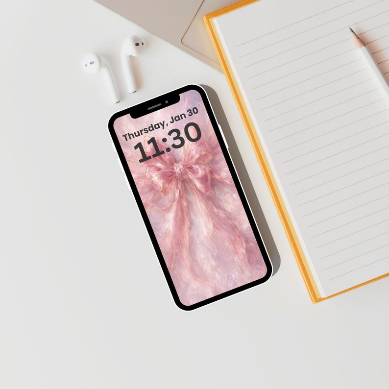 May include: A smartphone shows a pink bow wallpaper with the time "11:30" and the date "Thursday, Jan 30". White earbuds and a yellow-edged notebook with a pencil are on a white surface.