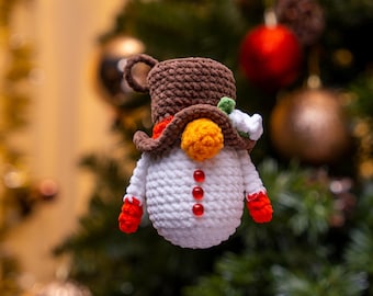 May include: A handmade crochet snowman Christmas ornament. The snowman has a brown top hat, orange nose, and red gloves. It has three red buttons down the front and is decorated with a small flower. The ornament is hanging in front of a Christmas tree.