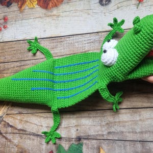 May include: A handmade green crochet alligator toy with blue stripes, white eyes, and a red tongue. The alligator is lying on a wooden surface with fall foliage and pumpkins in the background. The alligator's mouth is open.