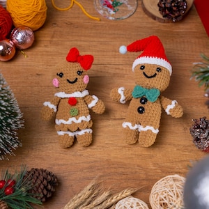 May include: Two handmade crochet gingerbread people. One has a red bow, the other a Santa hat. Both have white trim, smiling faces, and decorative buttons. Festive holiday decor on a wooden surface.
