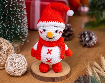 Puede incluir: Una figura de muñeco de nieve de ganchillo hecha a mano. El muñeco de nieve lleva un gorro, bufanda y mitones rojos, con una nariz de zanahoria y ojos de carbón. Se encuentra sobre una rodaja de madera, ideal para la decoración navideña.