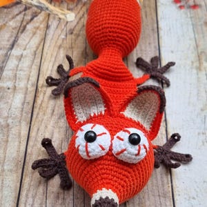 Op de afbeelding: Een handgemaakte, gehaakte vossenknuffel in fel oranje, met een wit en bruin getipte staart. De vos heeft grote, expressieve ogen met zwarte pupillen en rode details. De oren en poten van de vos zijn bruin. Op een houten ondergrond.