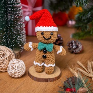 May include: A crocheted gingerbread man figurine wearing a red Santa hat with a white trim and a blue pom-pom. The gingerbread man has a green bow tie, black buttons, and white icing details. It is standing on a wooden slice, with Christmas decorations in the background.