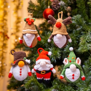 Könnte beinhalten: Fünf handgefertigte gehäkelte Weihnachtsschmuckstücke in Form von Wichteln, die an einem Weihnachtsbaum hängen. Die Wichtel haben verschiedene Hüte und Outfits in Rot, Braun, Weiß und Grün. Jedes Ornament ist etwa 10 cm hoch.