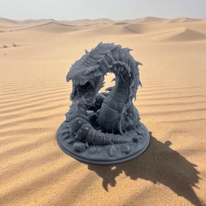 May include: A grey, 3D-printed miniature figurine of a segmented, serpentine creature with a wide, open mouth, sharp teeth, and numerous spikes along its body, coiled atop a circular base with textured, rocky details on the surface.