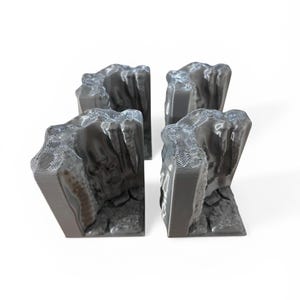 Op de afbeelding: Vier grijze, 3D-geprinte rotsformaties met getextureerde oppervlakken en gelaagde details. De formaties hebben een ruwe, rotsachtige uitstraling, met een basis die lijkt op gebroken steen. De objecten zijn geplaatst tegen een witte achtergrond.