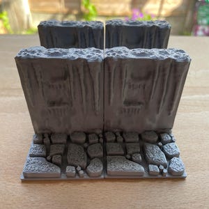 Op de afbeelding: Vier grijze, 3D-geprinte miniatuur terreinstukken. Elk stuk heeft een rotsachtige basis en een klifachtige structuur. De stukken zijn in een vierkante formatie gerangschikt, geschikt voor tabletop gaming of modelbouw.