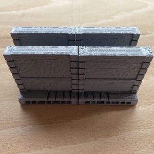 Op de afbeelding: Een grijze, 3D-geprinte miniatuur muursectie, bestaande uit vier verbonden stukken. De muur heeft een textuur, die lijkt op stenen blokken, en is ontworpen voor tabletop games of modelbouw. Het algehele ontwerp is rechthoekig.