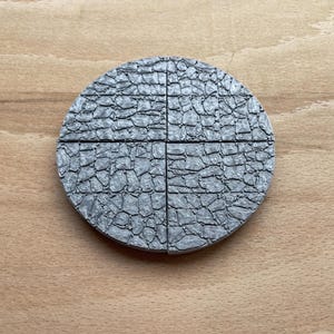 Op de afbeelding: Een cirkelvormig object met een grijze, steenachtige textuur, verdeeld in vier delen. Het oppervlak heeft een gedetailleerd, ruw uiterlijk, vergelijkbaar met kasseien. Op een lichtbruine houten achtergrond.