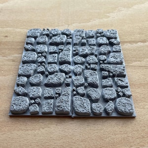 May include: Two grey rectangular panels featuring a stone wall design. The panels are covered in various sizes and shapes of stones, creating a realistic appearance. These panels are likely for tabletop gaming or model building, with a detailed stone effect.