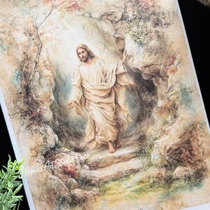 May include: A religious art print depicting a figure in flowing white robes emerging from a stone cave. The artwork features soft, muted colors, with a focus on the figure's face and outstretched hand. The background includes a rocky landscape and foliage.