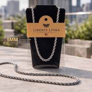 Può includere: Una collana a catena a corda color argento, spessa 5 mm, esposta su un espositore per gioielli in velluto nero. L'espositore ha un'etichetta marrone con il testo "LIBERTY LINKS - AMERICAN BESTICA" e un logo a forma di aquila. La collana è mostrata su uno sfondo urbano sfocato.