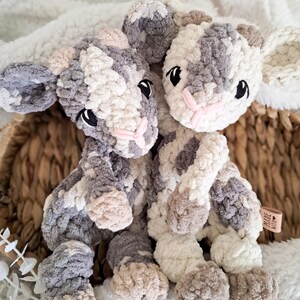 May include: Two handmade crochet stuffed animal toys. The toys are in the shape of a calf, with a mix of gray, white, and tan yarn. They have black eyes and pink noses. The toys are sitting on a white, fluffy surface.