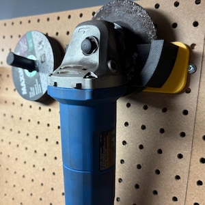 May include: A blue and grey angle grinder with a black and yellow guard is mounted on a pegboard. A stack of grinding discs is to the left. The tool is designed for cutting and grinding.