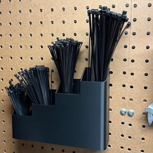 Puede incluir: Un organizador negro escalonado lleno de bridas negras está montado en una panel perforado. Una llave inglesa plateada y otras herramientas cuelgan cerca. El panel perforado es de madera.