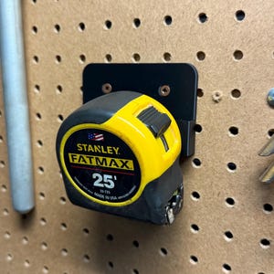 Puede incluir: Un flexómetro Stanley FatMax amarillo y negro de 7,62 metros, montado en un soporte negro. El flexómetro está fijado a una tabla perforada, con unos alicates a la derecha.