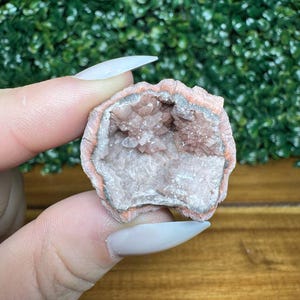 Puede incluir: Una geoda con un exterior rosa anaranjado y un grupo de cristales rosa claro en el interior. La geoda se sostiene entre los dedos sobre un fondo verde. Los cristales son visibles en el centro de la geoda.