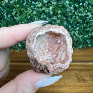 Puede incluir: Una peque&ntilde;a geoda redonda con un exterior &aacute;spero de color marr&oacute;n rojizo y un interior cristalino rosado. La geoda se sostiene en una mano, mostrando su textura natural y variaciones de color. La geoda es una piedra natural.
