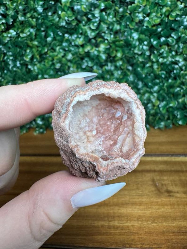 Puede incluir: Una peque&ntilde;a geoda redonda con un exterior &aacute;spero de color marr&oacute;n rojizo y un interior cristalino rosado. La geoda se sostiene en una mano, mostrando su textura natural y variaciones de color. La geoda es una piedra natural.