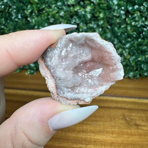 Puede incluir: Geoda de color rosa claro con interior cristalino. La geoda tiene un exterior rugoso y texturizado con una banda blanca y rosa. La geoda se sostiene entre dos dedos.