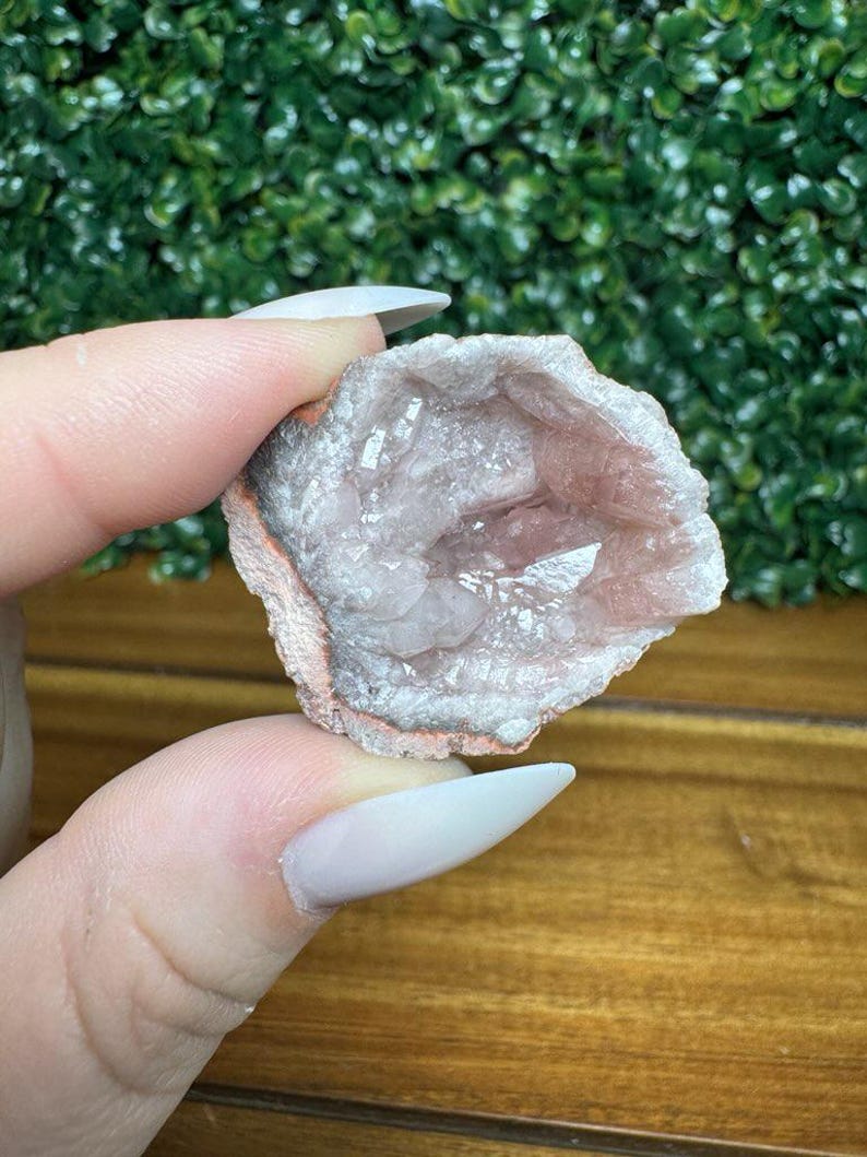 Puede incluir: Geoda de color rosa claro con interior cristalino. La geoda tiene un exterior rugoso y texturizado con una banda blanca y rosa. La geoda se sostiene entre dos dedos.