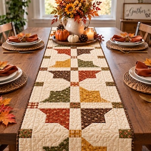 May include: A rectangular quilted table runner featuring a repeating star pattern in autumn colors including orange, brown, yellow, and green on a cream background. The runner is centered on a wooden table with place settings and a floral centerpiece.