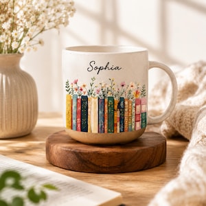 May include: A speckled off-white ceramic mug featuring the name Sophia in black script above a colorful illustration of a row of books with wildflowers growing from the tops. The mug has a sturdy handle and a smooth, rounded base design.