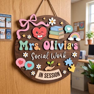 May include: A round wooden door sign featuring the text Mrs. Olivia's Social Work and IN SESSION. The sign is decorated with colourful 3D cutouts including a winking heart, a stack of books, a rainbow, flowers, stars, speech bubbles, and a hand holding a sprout.