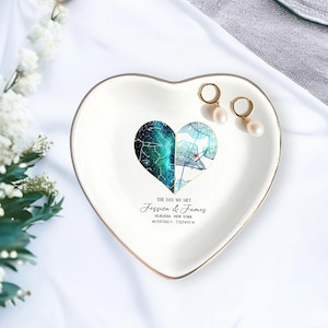 May include: Heart-shaped white ceramic trinket dish with a gold rim. The dish features a split heart design with a starry night sky and a city map. Text reads "THE DAY WE MET" and names, date, and coordinates. Two gold hoop earrings with pearls rest on the dish.