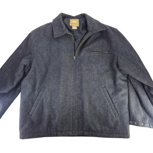 Puede incluir: Chaqueta de lana gris oscuro con cierre de cremallera completo, cuello y dos bolsillos delanteros. La chaqueta tiene un pequeño bolsillo en el pecho y detalles de botones en los puños. La etiqueta de la marca es visible en el interior del cuello.