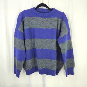May include: A blue and gray striped sweater hangs on a black hanger. The sweater has a crew neck and long sleeves. The stripes are horizontal and alternate between a vibrant blue and a muted gray. The sweater is made of a soft, knitted material.