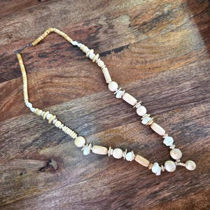 May include: A necklace featuring a variety of light beige and off-white beads in different shapes and sizes, including cylindrical, spherical, and irregular disc-like forms, strung together on a thin cord with a visible metal clasp at the end.