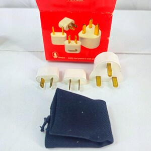 Könnte beinhalten: Ein Set weißer Reiseadapter mit goldenen Stiften, präsentiert mit einer roten Box und einem marineblauen Kordelzugbeutel. Auf der Box ist der Markenname "FRANZUS" aufgedruckt.