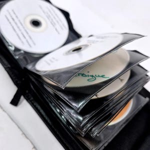 May include: A black CD binder with multiple white CDs inside. The CDs are in clear plastic sleeves. Some CDs have writing on them. The binder is zippered and has a black fabric exterior.
