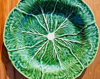 May include: A decorative ceramic plate with a cabbage leaf design. The plate is a vibrant green with white veins and a white center. The plate is on a wooden surface.