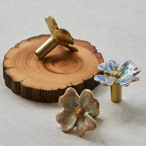 May include: Three flower-shaped cabinet knobs. One is on a wooden slice, and the other two are on a light gray surface. The knobs are gold-colored with pastel-colored enamel petals and a gold center. The knobs are likely for furniture.