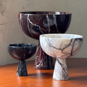 May include: Three decorative marble bowls with pedestal bases. The bowls have a rounded top and are in different sizes. The marble has a dark red, black, and white veining pattern. The bowls are displayed on a wooden surface.