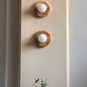 May include: Two round wall sconces with a marble-like appearance, each featuring a central white globe light. The sconces are mounted on a light beige wall. A wooden vase with green foliage sits below.