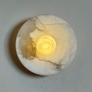 May include: A round, marble-like wall sconce with a central, glowing, yellow light. The sconce has a circular shape with a marbled, off-white and gray pattern. The light source is a smaller, spherical shape in the center, emitting a warm, yellow glow.