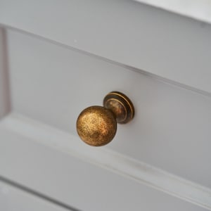 May include: A round cabinet knob with a textured, antique brass finish featuring dark brown and gold mottled tones. The knob is mounted on a light grey drawer front with a smooth, matte surface and a horizontal recessed panel detail.