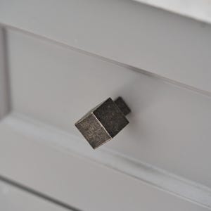 May include: A square-shaped cabinet knob with a dark, weathered metallic finish. The knob features a textured, aged surface and is mounted onto a light gray drawer front with a recessed panel design and a white marble countertop edge above.
