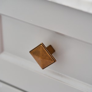 Op de afbeelding: Een vierkante kastknop met een metallic bronzen afwerking en een ontwerp met afgeschuinde randen. De knop is gemonteerd op een platte, lichtgrijze ladefront en is voorzien van een centrale pin die het vierkante vlak met de lade verbindt.